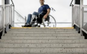 Imagem mostra um rapaz cadeirante usando camiseta azul marinho e calça jeans. Ele está no topo de uma escada de concreto olhando para baixo pensativo. Seu rosto está desfocado. A imagem toda puxa para o cinza claro